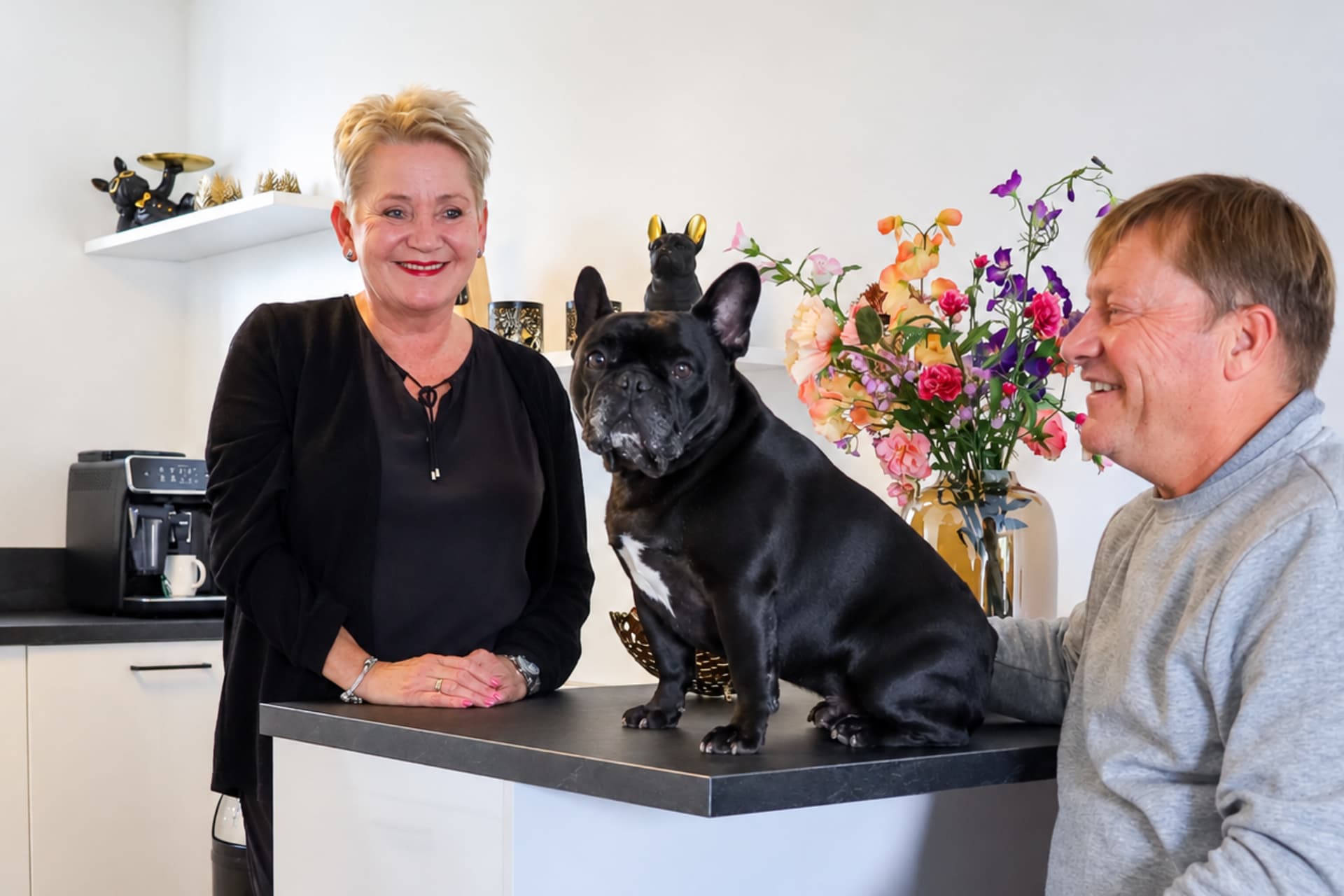 Binnen kijker Dick en Ingrid.png Witte keuken met zwart werkblad waar een Franse Bulldog op zit en een ouder stel omheen staat
