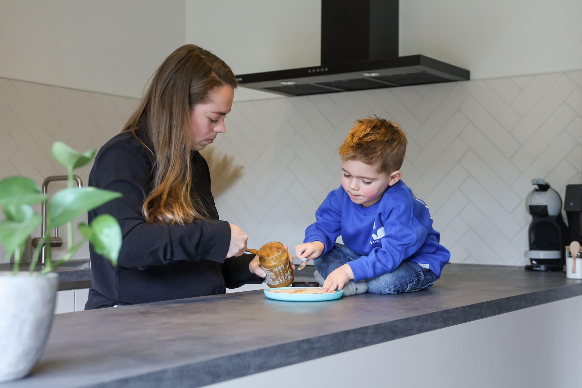 Binnen kijker Richard en Daisy.png Witte keuken met grijs werkblad waar een jong jongetje op zit die met zijn moeder eten bereid